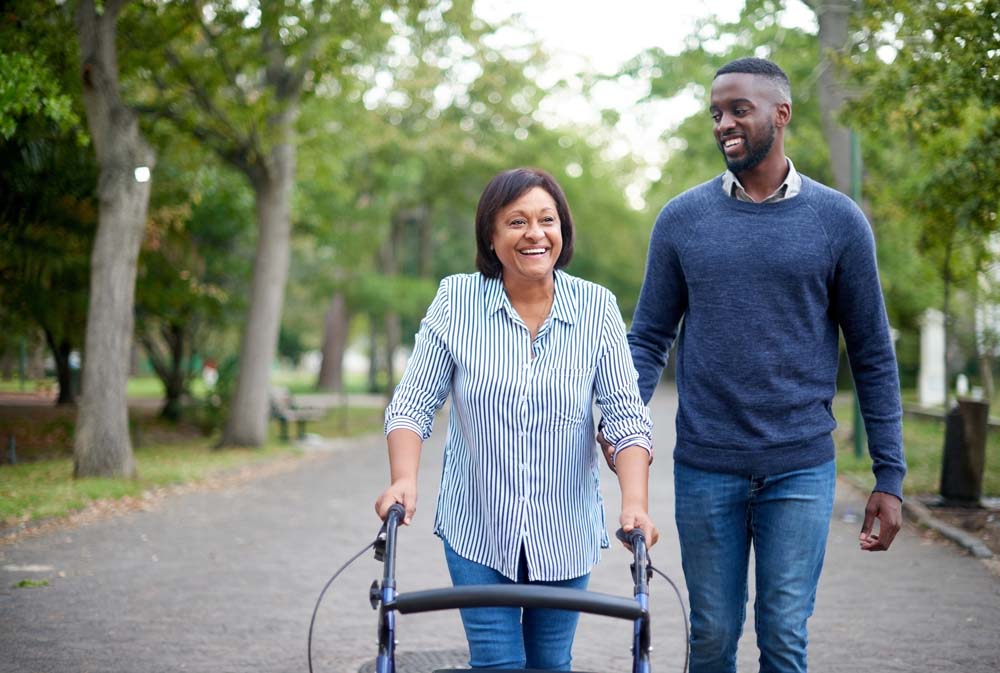 Woman with mobility issues walking with walking frame and assisted by carer.